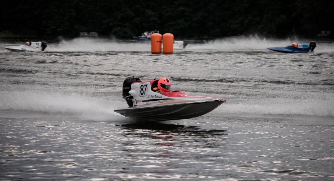 Zdjęcie przedstawia sportową motorówkę z numerem 87 mknącą po tafli jeziora. Sternik ubrany w czerwony kask. Zdjęcie wykonane podczas zawodów. Na wodzie rozieszczone elementy wyznaczające trasę.