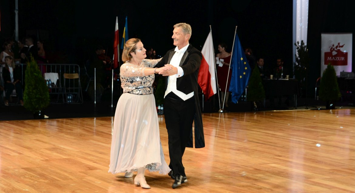 Para tanczena podczas Mistrzostw Polski w Tańcach Polkich "Mazur" na parkiecie hali sportowej w Wieliszewie. Kobieta w kremowej sukni, mężczyzna we fraku. W tle stojak z flagami Polski i UE.