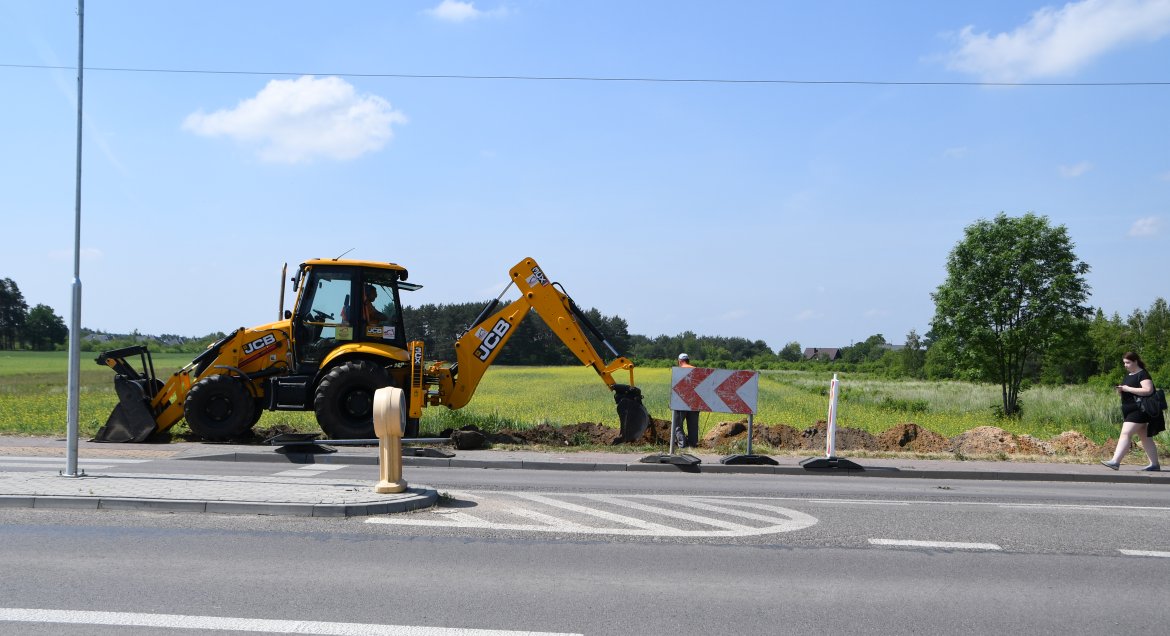 Zdjęcie, na pierwszym planie jezdnia asfaltowa, po lewej latarnia, wyspa separacyjna i chodnik. Dalej pracująca, żółta koparka. Po prawej stronie zdjęcia idąca chodnikiem kobieta.