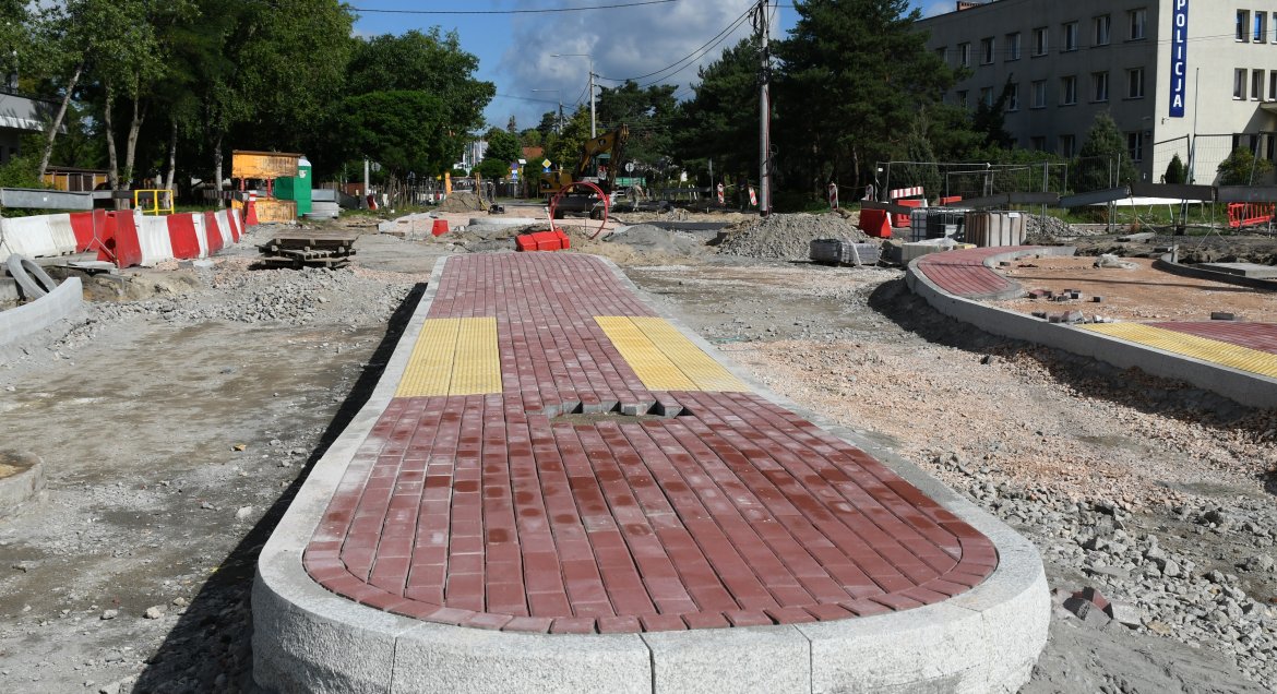 Zdjęcie: prace budowlane na skrzyżowaniu ulic Jagiellońskiej i Mickiewicza w Legionowie. Powstaje rondo. Na pierwszym planie, centralnie, wyspa separacyjna z czerwonej kostki betonowej, po obu jej stronach wydołowane miejsce na jezdnie wysypane kruszywem. W tle drzewa, po prawej budynek z szyledem policja.