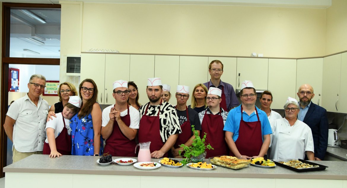 Zdjęcie, grupa szesnastu osób, w tym osoby z niepełnosprawnościami, osób pozuje do zdjęcia w kuchni w Powiatowym Zespole Szkół i Placówek Specjalnych w Legionowie. Na stole przed nimi stoja potrawy.