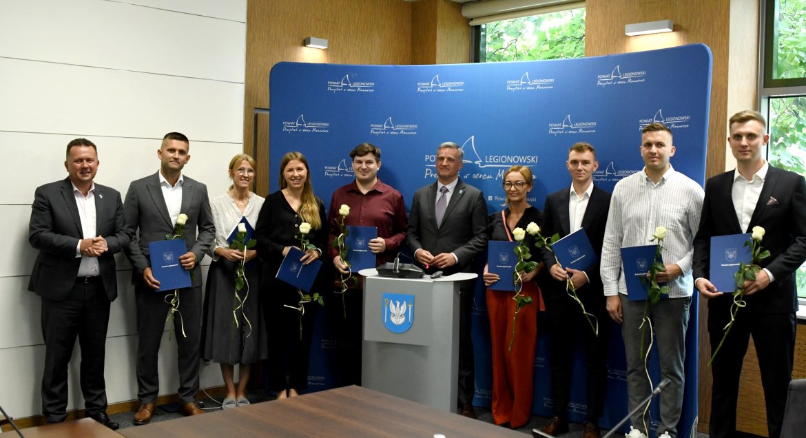 Zdjęcie wykonane podczas sesji Rady Powiatu w sali konferencyjnej legionowskiego ratusza. Grupa osób pozuje na tle ścianki promocyjnej Powiatu Legionowskiego. Osoby trzymają w rekach dyplomy oraz róże.