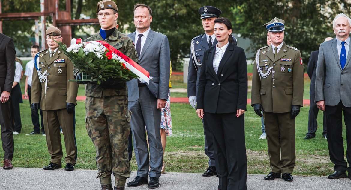 Zdjęcie: na pierwszym planie w centralnym punkcie stoi żołnierz trzymający wieniec z biało-czerwoną szargą, na drugim planie stoją przedstawiciele wojska, władz samorzadowych i inni zaproszeni goście.