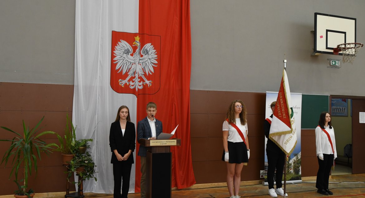 Rozpoczecie roku szkolnego 2024-2025 w Liceium Ogólnokształaczacym im. Marii Konopnickiej w Legionowie. Wydarzenie odbywa sie na sali gimnastycznej. Na zdjeciu ubrana na galowo młodzież w poczcie sztandarowym. Jedna osoba czyta przy mównicy.