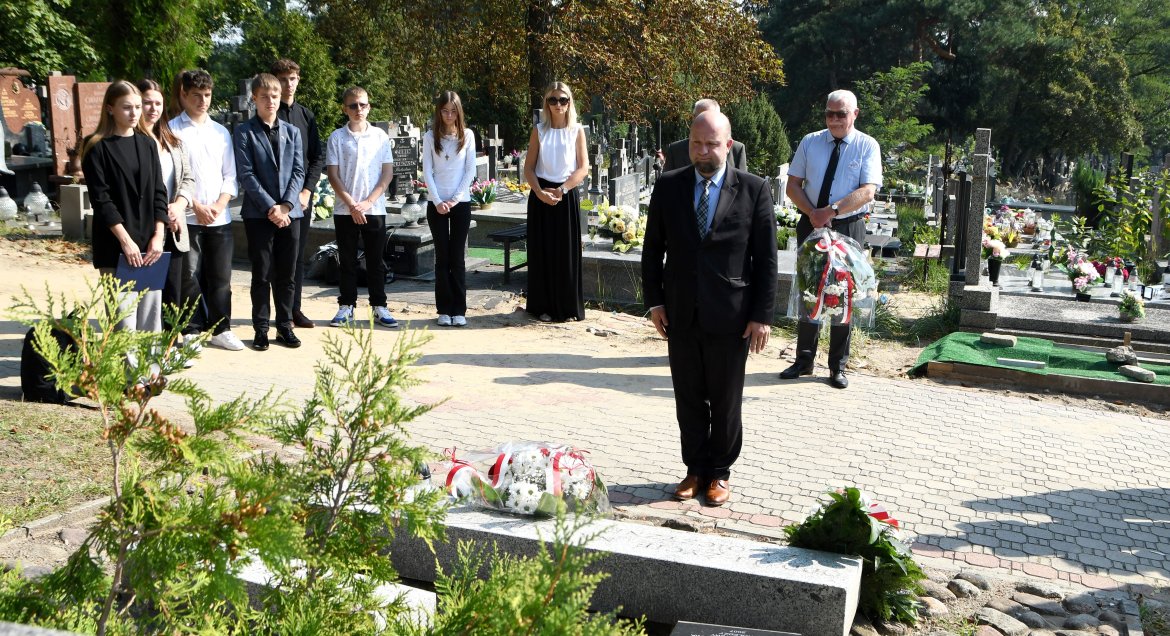 Zdjęcie wykonane na cmentarzu w Legionowie. Na pierwszym planie mężczyzna oddający hołd pod pomnikiem  „Oddali życie na nieludzkiej ziemi za Ojczyznę i swoje miasto”. Na drugim plenie grupa jedenastu osób, głównie młodzieży licealnej. W tle nagrobki.