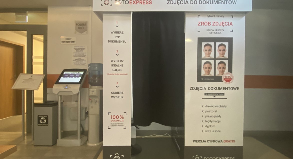 Zdjęcie przedstawia automat do robienie zdjęć. Biała budka z napisami informacyjnymi, wejście zasłonięte czarną kotarą.