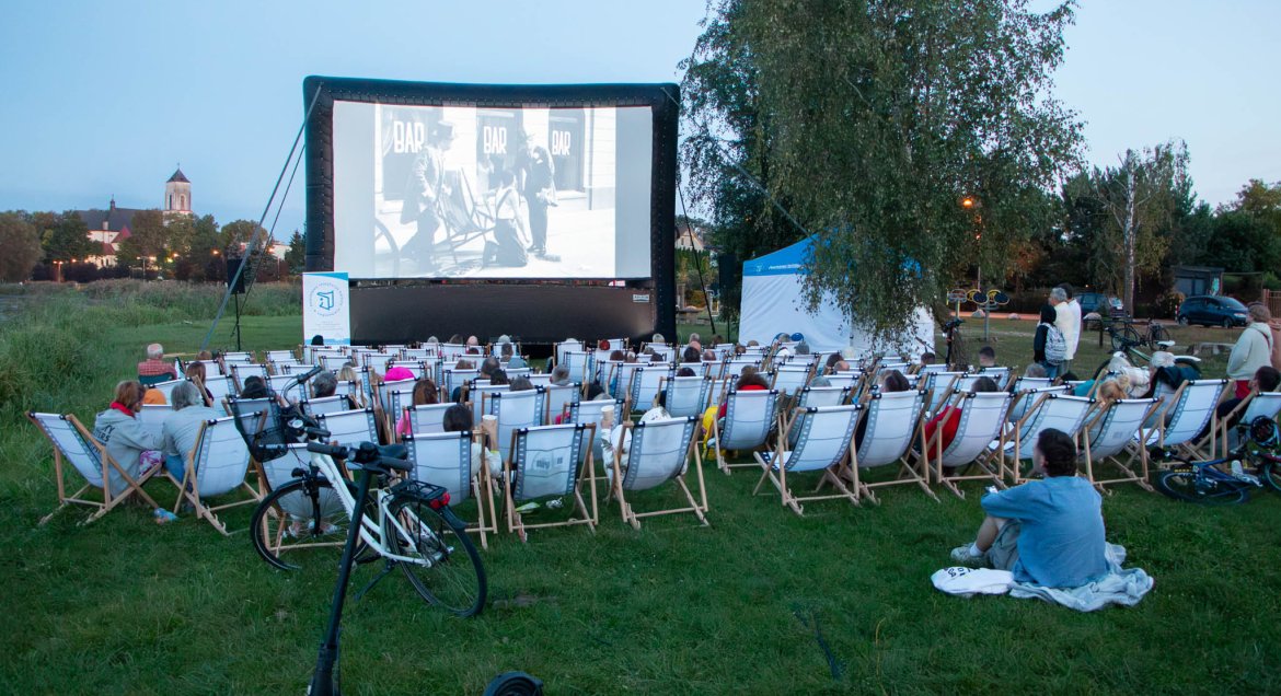 Zdjecie plenerowe: osoby siedzące na leżakach oglądają film wyświetlany na duzym ekranie.