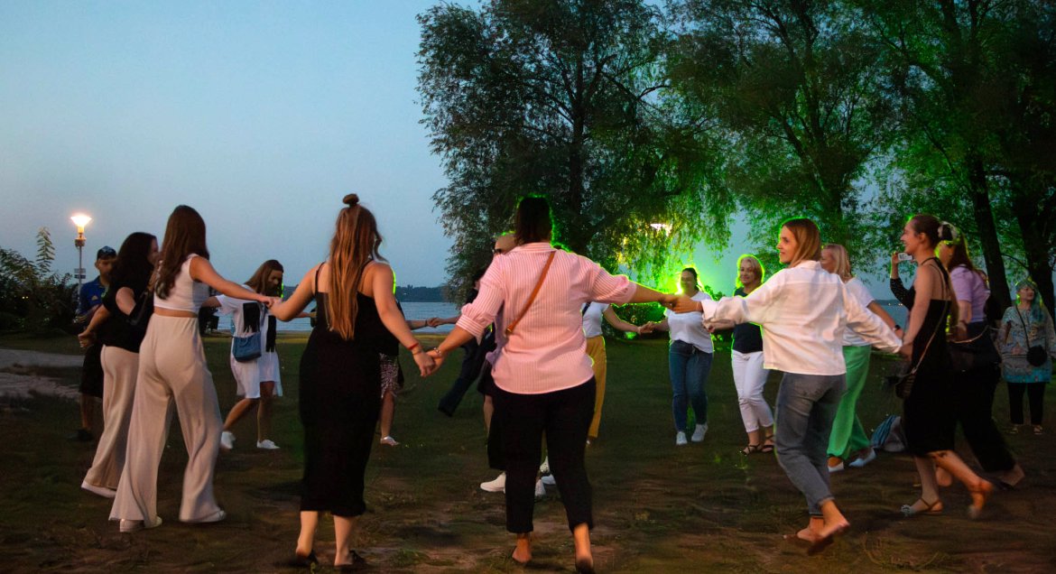 Wieczorne zdjęcie: osoby tańczace w okręgu na plazy nad Jeziorem Zegrzyńskim.