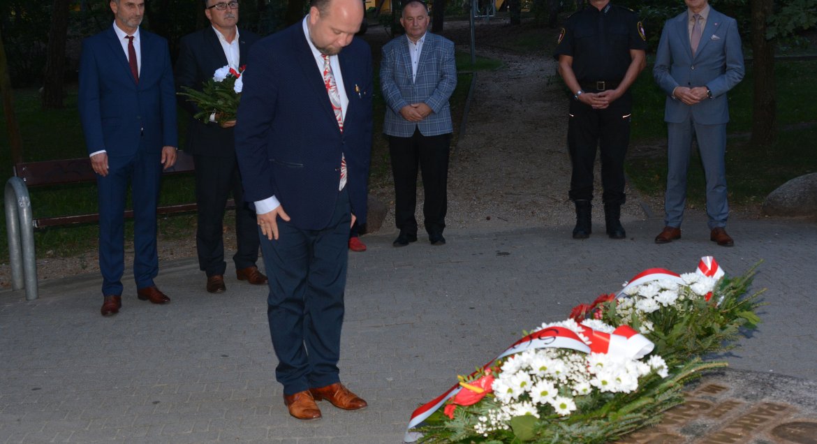 Zdjęcie: w centralnym punkcie mężczyzna w garniturze oddaje hołd przy obelisku pamiątkowym znajdującym się w Parku im. NSZZ „Solidarność” w Legionowie. W tle pięciu mężczyzn, uczestników uroczystości.