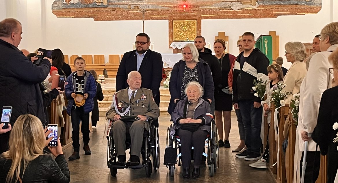 Zdjęcie wykonane w kościele pw. Najświętszego Ciała i Krwi Chrystusa w Legionowie. W tle ołtarz, po bokach ławki z wiernymi, po srodku dwóch weteranów na wózkach inwalidzkich prowadzeni przez opiekunów.