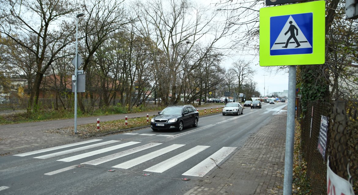 Zdjęcie przedstawia przejście dla pieszych przy skrzyzowaniu ulic Sobieskiego i Skargi w Legionowie. Po prawej na pierwszym planie znak ostrzegawczy, żólte tło + piktogram dziecka. Na kolejnym planie pasy na jezdni, w tle budynki mieszkalne.