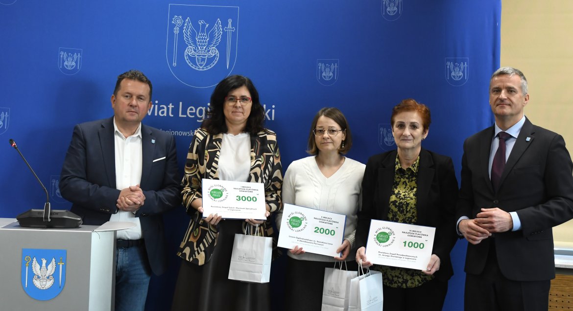 Zdjęcie: trzy kobiety (w środku) oraz dwóch mężczyzn (po bokach) pozują na tle ścianki reklamowej Powiatu Legoonowskiego. Kobiety trzymają w rękach tabliczki z voucherami. Wręczenie nagród w konkursie ekologicznym.