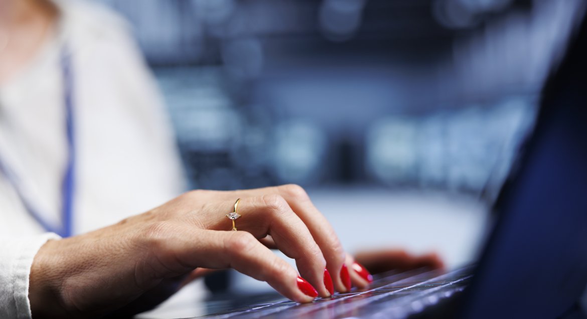 Na zdjęciu widać damską dłoń piszącą na klawiaturze komputera. Na jednym z jej palców znajduje się pierścionek z białym kamieniem, a paznokcie pomalowane są na kolor czerwony.