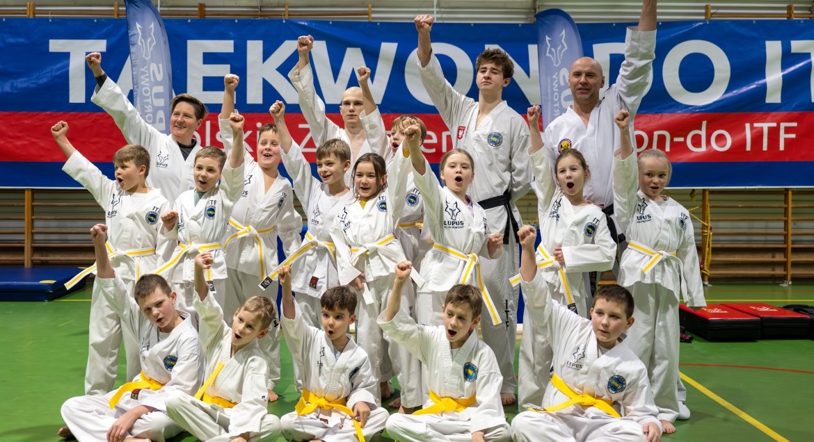Nas zdjeciu dziecięcy zawodnicy taekwondo, z trenerami, dzieci unoszą ręce w górę, ubrane są w białe kimona