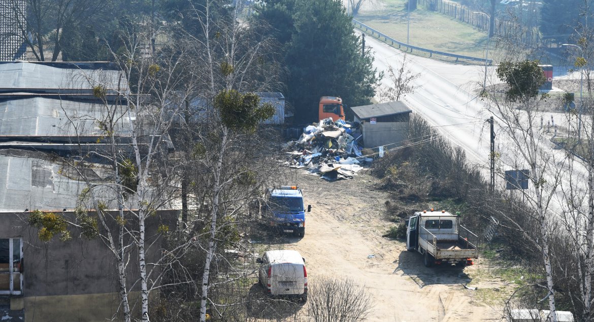 Widok z góry - po lewej stronie: trzy budynki do rozbiórki; po prawej stronie nieutwardzona droga oraz samochody dostawcze. Po obu stronach zieleń.