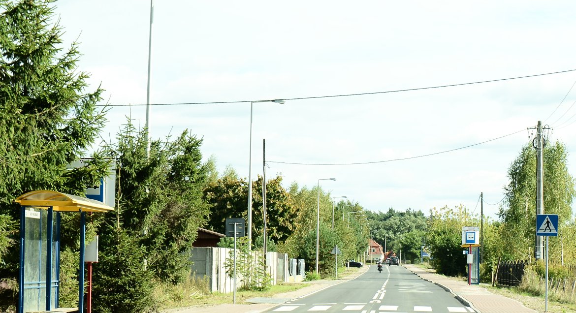 Zdjęcie: fragment drogi powiatowej nr 1810W w Gminie NIeporet, widok na asfalt, przejście dla pieszych oraz przystanek autobusowy. Po obu stronach roślinność oraz zabudowania.
