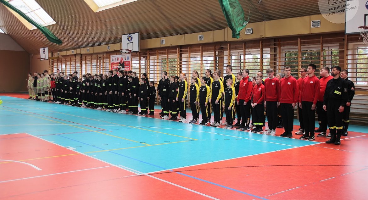 Młodzież w uniformach strażackich ustawiona w szeregu wzdłuż boiska w hali sportowej.