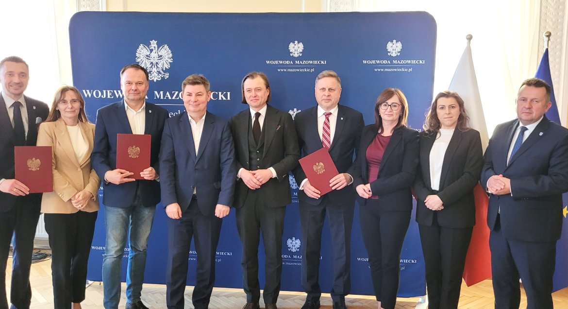 Grupa przedstawicieli władz samorządowych z terenu Mazowsze podczas pamiątkowego zdjęcia, pozują na granatowej ściance promocyjnej Wojewody Mazowieckiego.