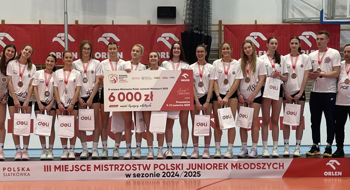 na zdjęciu drużyna siatkarek na podium, koło nich czek na 6 000 zł, na szyi medale, w rękach torby