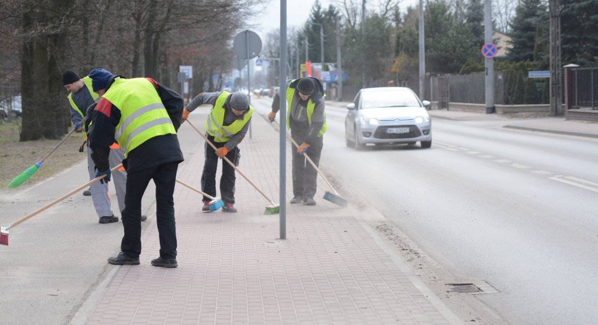 Pracownicy ubrani w odblaskowe kamizelki podczas prac porządkowych na poboczu drogi.
