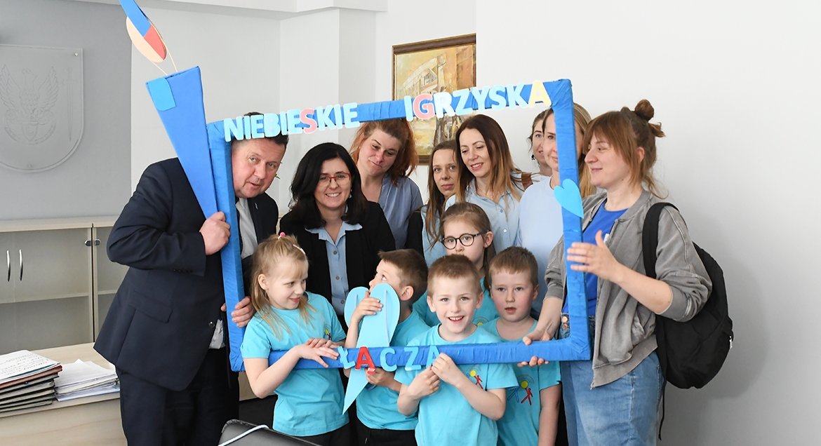 Grupa osób - przedszkolaków oraz dorosłych - pozująca w niebieskiej ramce z napisem: Niebieskie Igrzyska Łączą