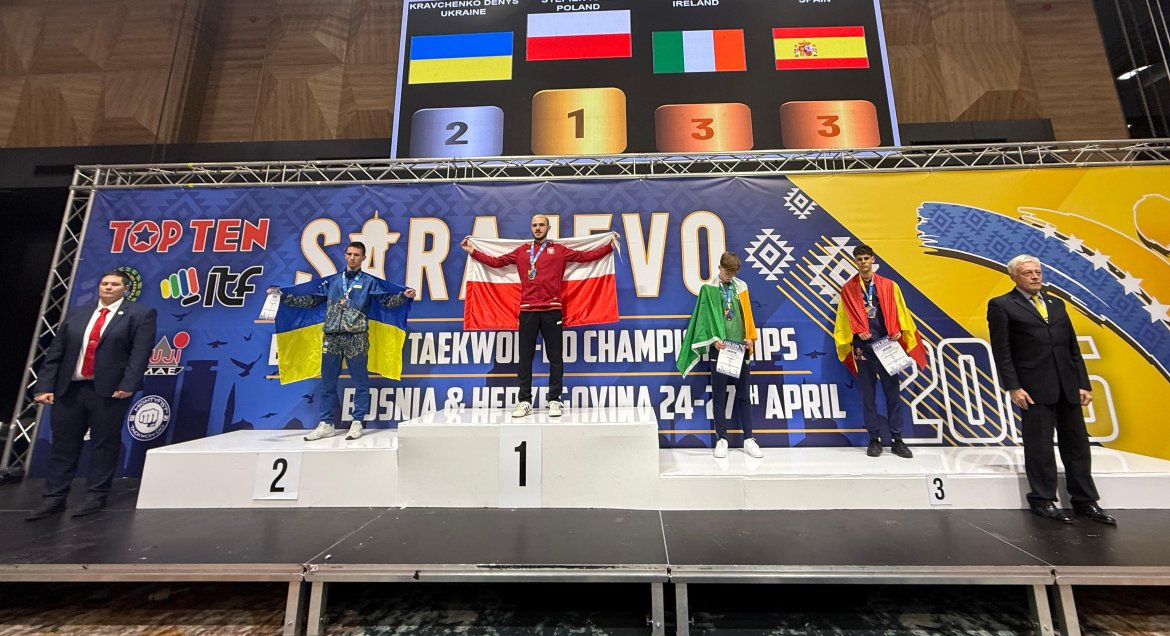 Igor Stępień na pidium w rękach sa sobą rozpięta flaga Polski, koło podium stoją dwaj mężczyźni