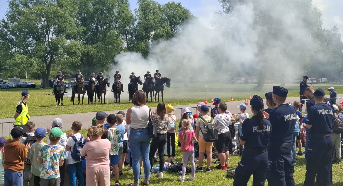 Plac, zgoromadzeni ludzie przed barierkami, za nimi policjanci na koniach robią pokaz