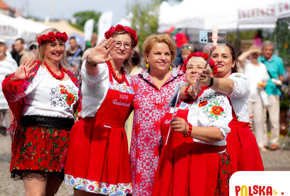 Na zdjęciu widać grupę uśmiechniętych kobiet w tradycyjnych strojach ludowych, pozujących do wspólnego selfie podczas plenerowego wydarzenia kulinarnego. Panie mają czerwone spódnice, kwieciste bluzki i ozdobne wianki na głowach, a w tle widoczne są białe namioty festiwalowe oraz zgromadzeni uczestnicy. W prawym dolnym rogu widnieje logo „Polska Od Kuchni – Festiwal Kulinarny”.