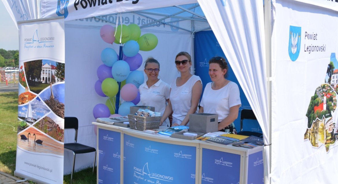Zdjęcie stoika promocyjnego Powiatu Legionowskiego, ww rodku trzy osoby przy ladzie