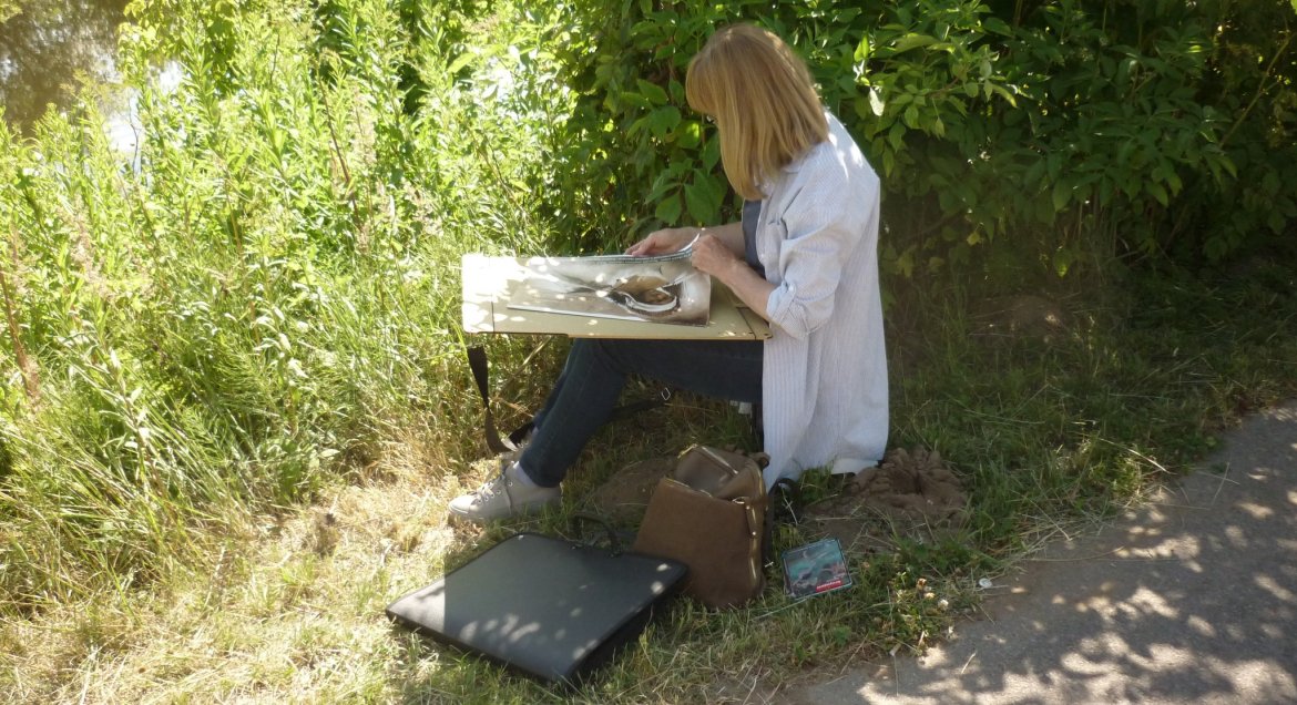 Na zdjęciu kobieta w otoczeniu zieleni przyrody, malująca obraz