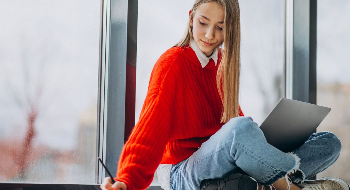 Na zdjęciu nastolatka siedząca na podłodze z laptopem na kolanach, robiąca notatki