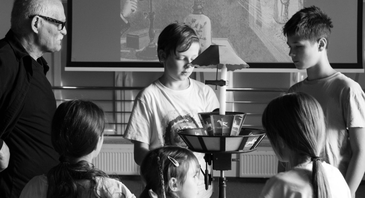 Czarnobiała fotografia przedstawiająca uczestników, którzy mogli przekonać się, jak funkcjonuje praksinoskop, czyli urządzenie w formie bębna obrotowego, w którym znajduje się taśma filmowa, co umożliwia widzenie ruchomego obrazu