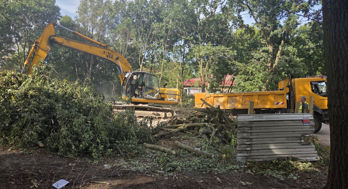 Zdjęcie przedstawia koparkę podczas prac rozbiórkowych na zadrzewionym terenie.