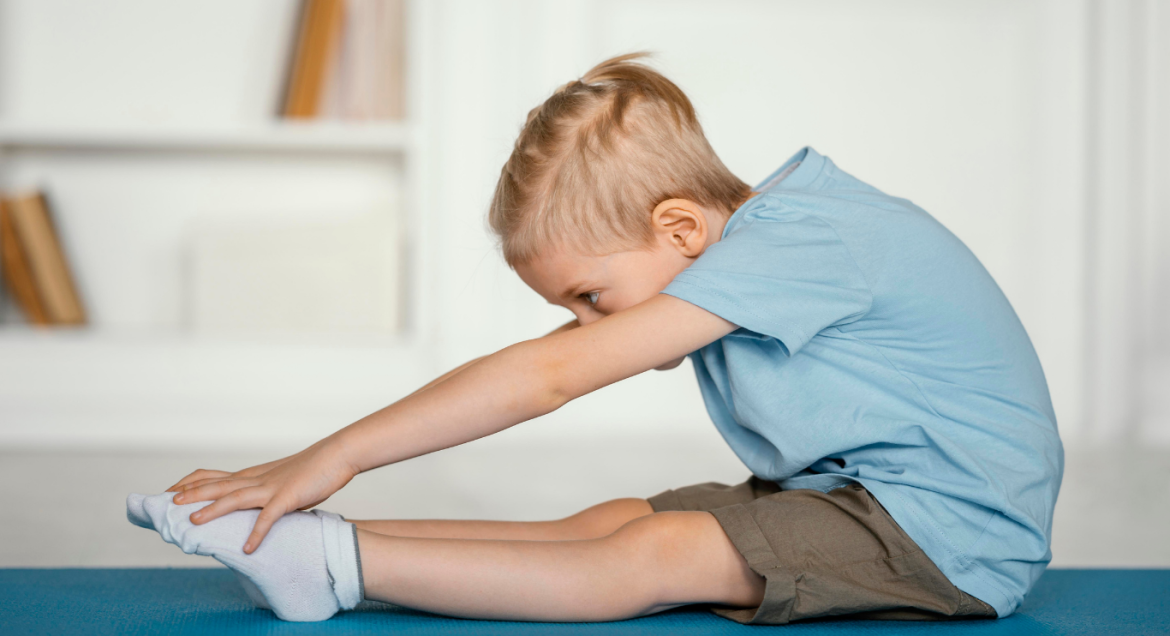 Na zdjęciu widać chłopca, który siedzi na podłodze z wyprostowanymi nogami i wykonuje skłon