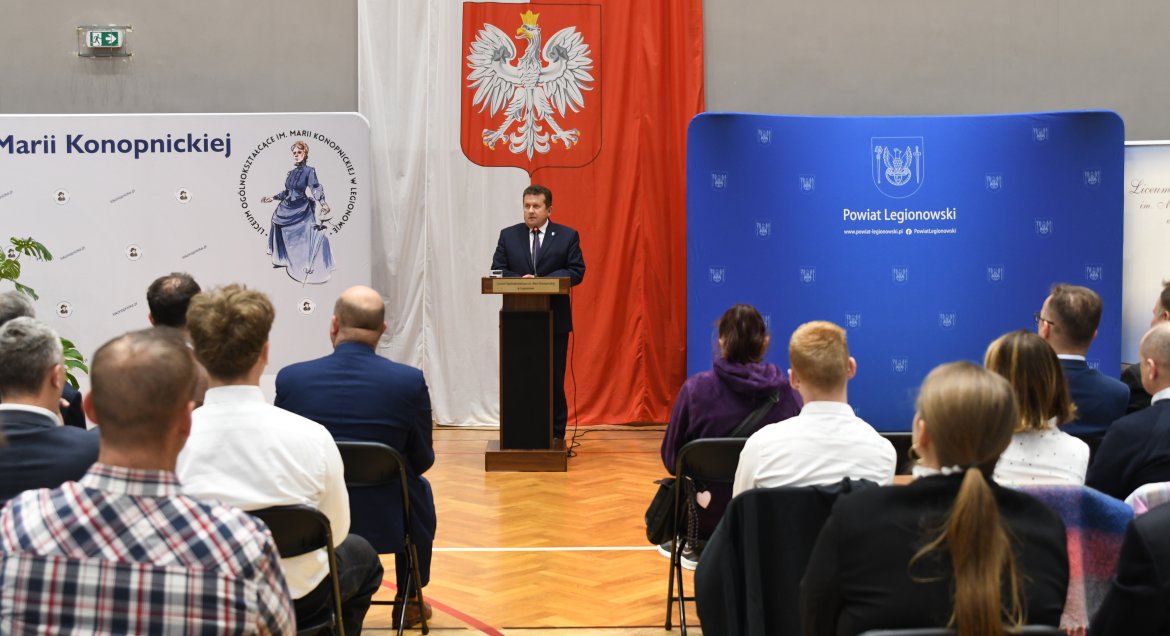 Zdjęcie wykonane na sali gimnastycznej LO im. Konopnickiej w Legionowie. Na pierwszym planie, tylem, zebrana publicznośc, w tle przy mównicy mężczyzna, za nim flaga państwowa oraz ścianki promocyjne.