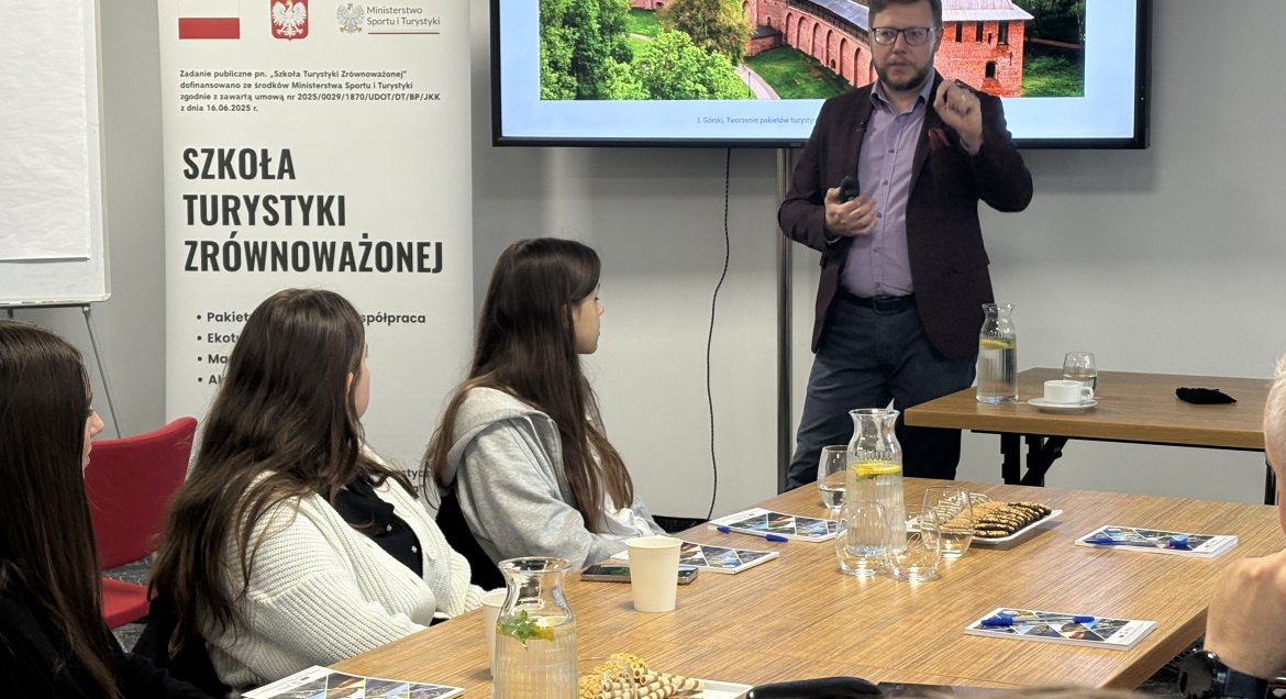 Uczestnicy szkolenia „Szkoła Turystyki Zrównoważonej” siedzą przy stołach konferencyjnych. Na pierwszym planie widoczna jest kobieta notująca w zeszycie, w tle trener omawia zagadnienia, korzystając z rzutnika.