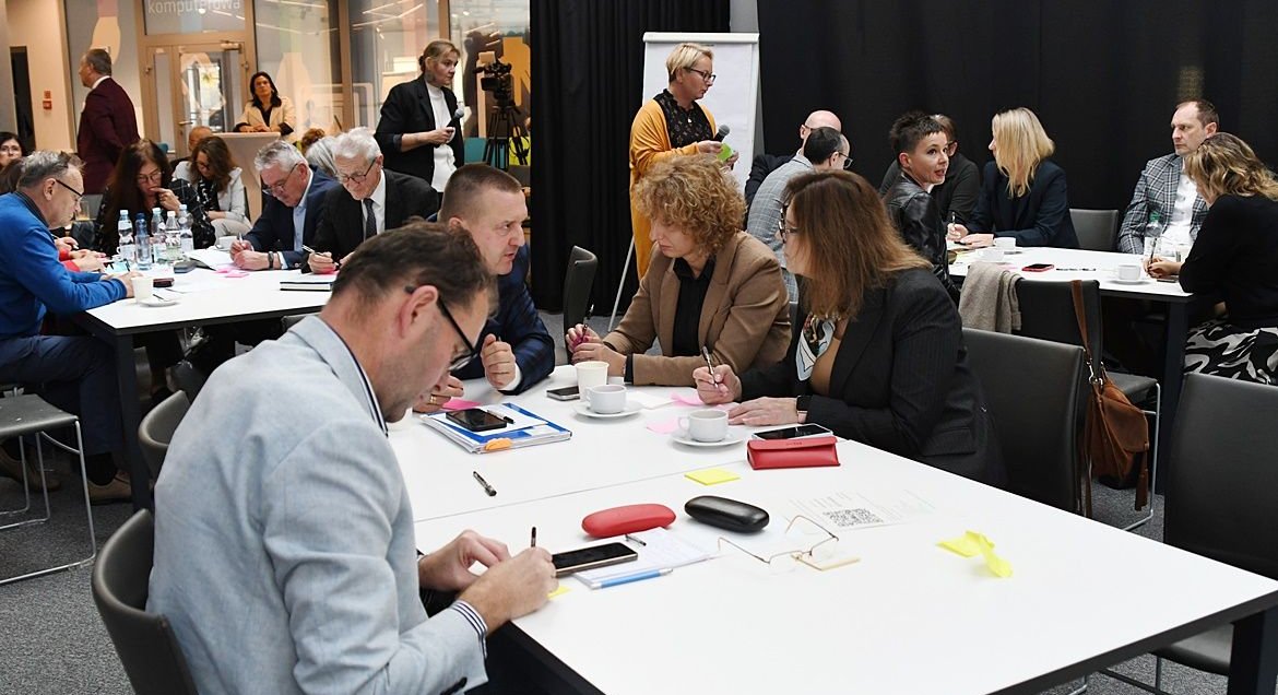 Na zdjęciu osoby siedzące przy stoliku. W tle widać więcej osób