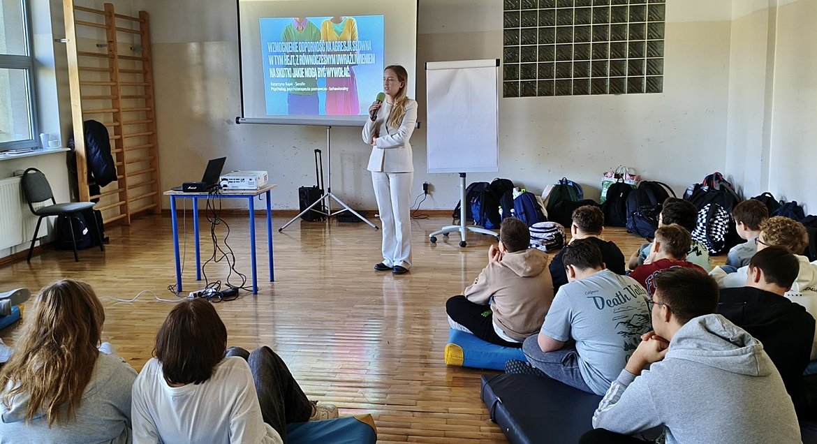 Grupa uczniów siedzi na sali. Na drugim planie osoba prowadzaca zajęcia.