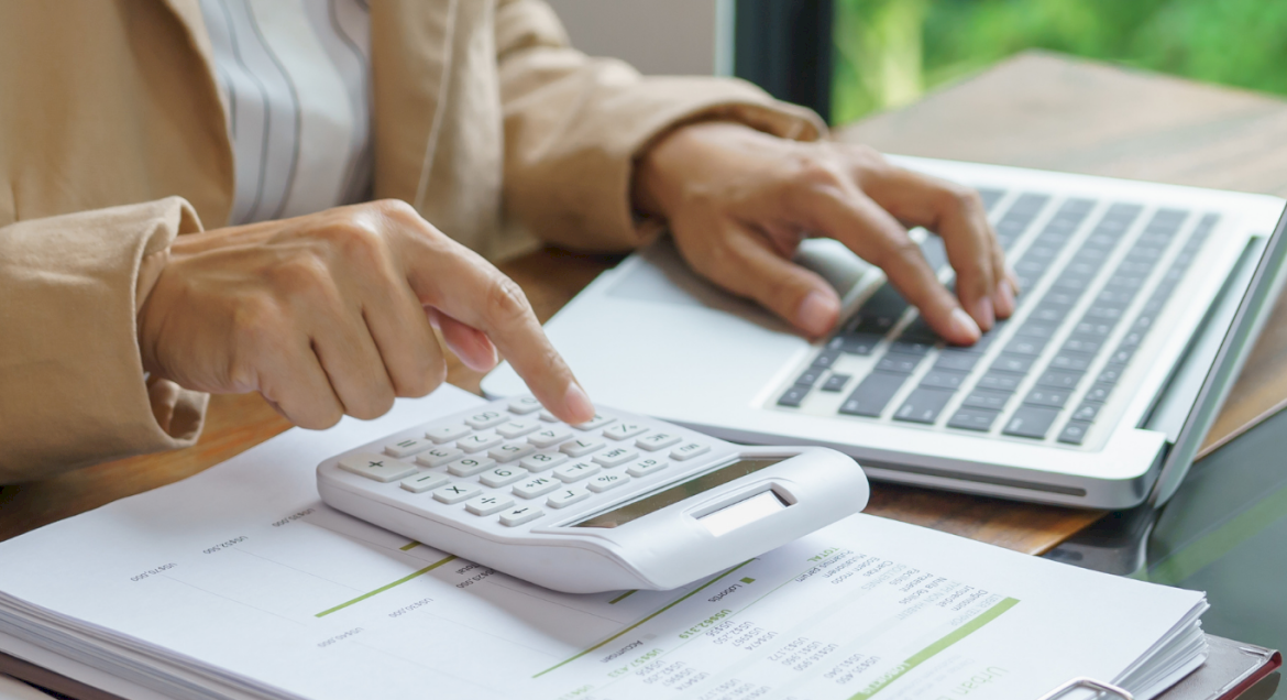 Na zdjęciu widać ręce kobiety, która liczy coś na kalkulatorze i ma przed sobą otwarty laptop