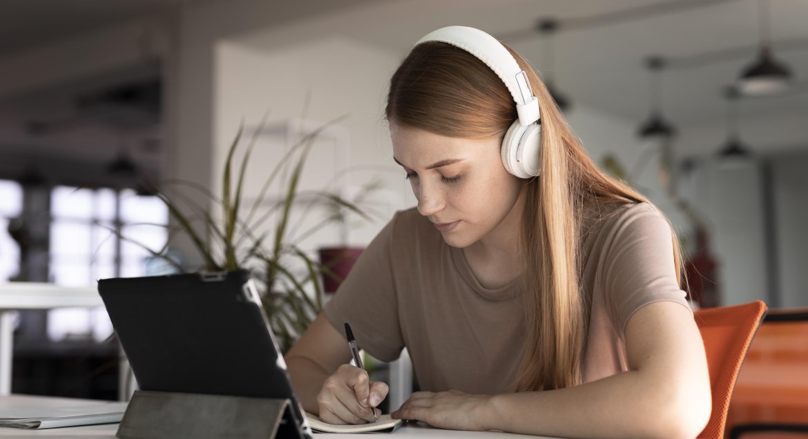 Zdjęcie: siedzaca przed tablektem młoda kobieta w słuchawkach.