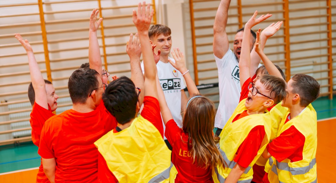 Na zdjęciu grupa młodzieży, ubrana w odblaskowe kamizelki, zebrana w koło i trzymająca uniesione wysoko prawe dłonie