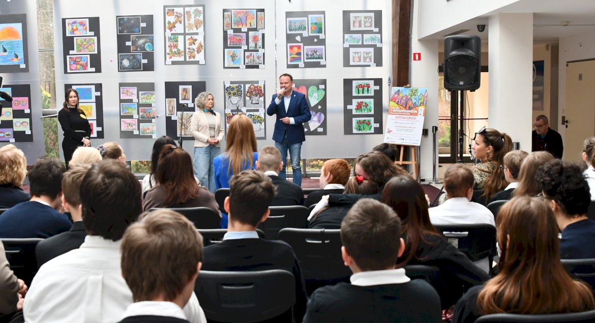 Pierwszy plan, widownia, dalej osoby przemawiające, w tle na ścianie wiszą prace będące częścią wystawy.