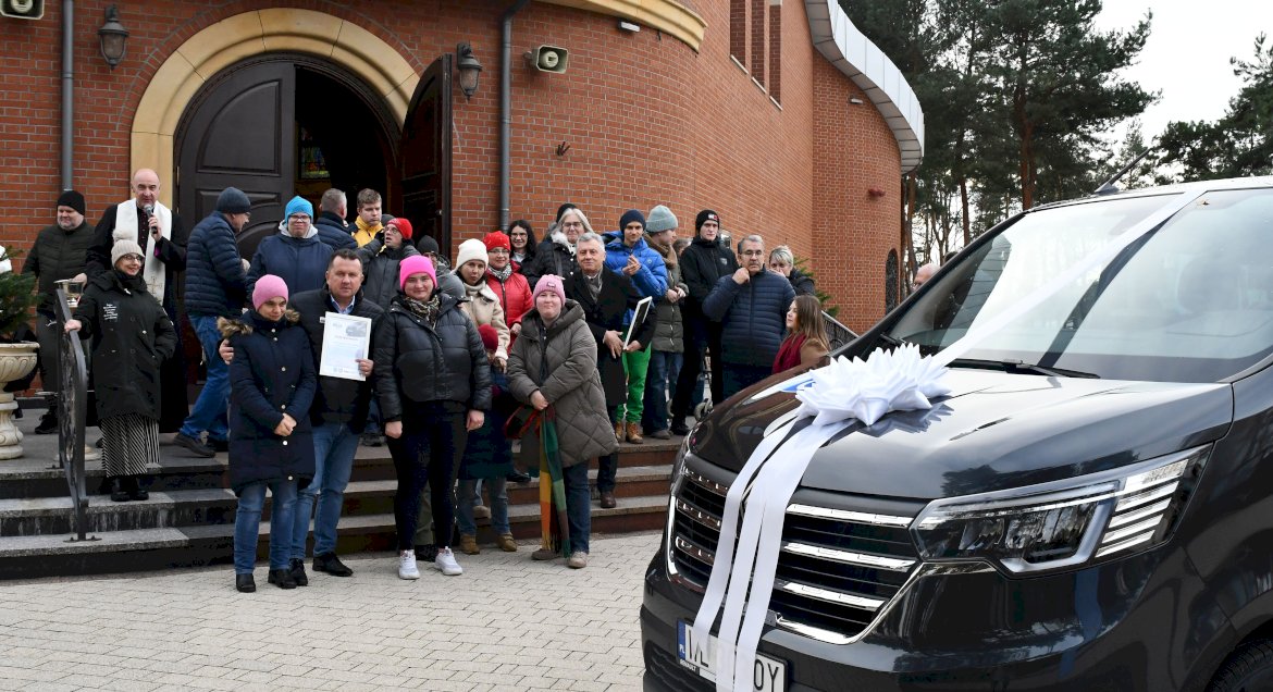 Zdjęcie: na pierwszym planie maska mikrobusu z kokardą na dugim planie sotjąca na schodach grupa osób, w tle kościół.