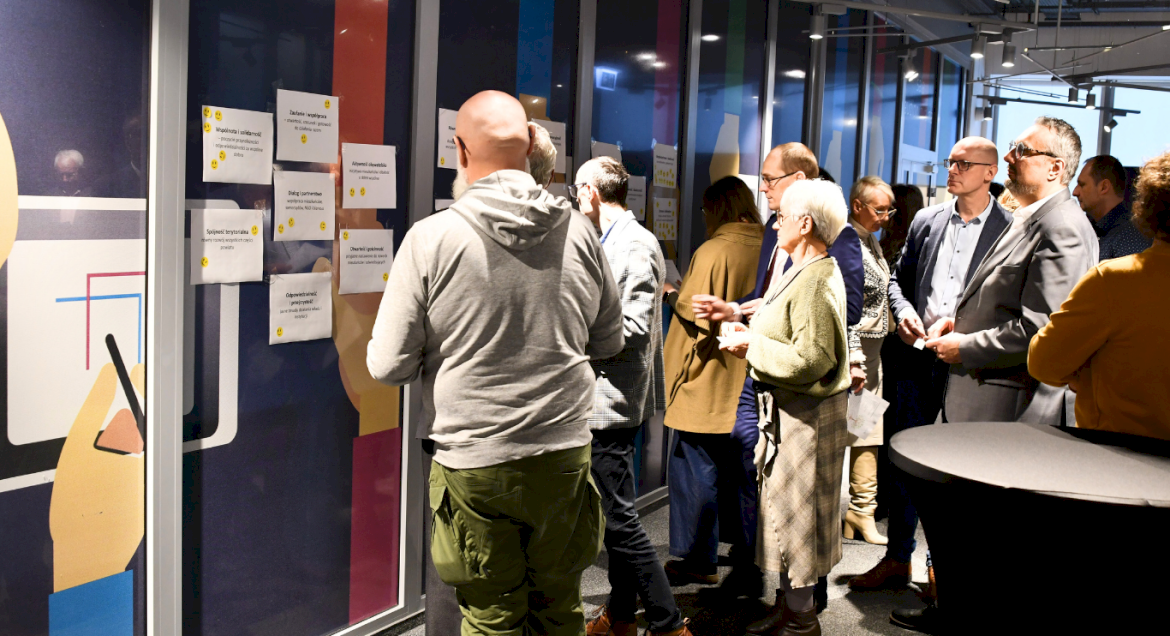 Na zdjęciu widoczni są członkowie warsztatów, którzy stoją przy ścianie za zawieszonymi kartkami