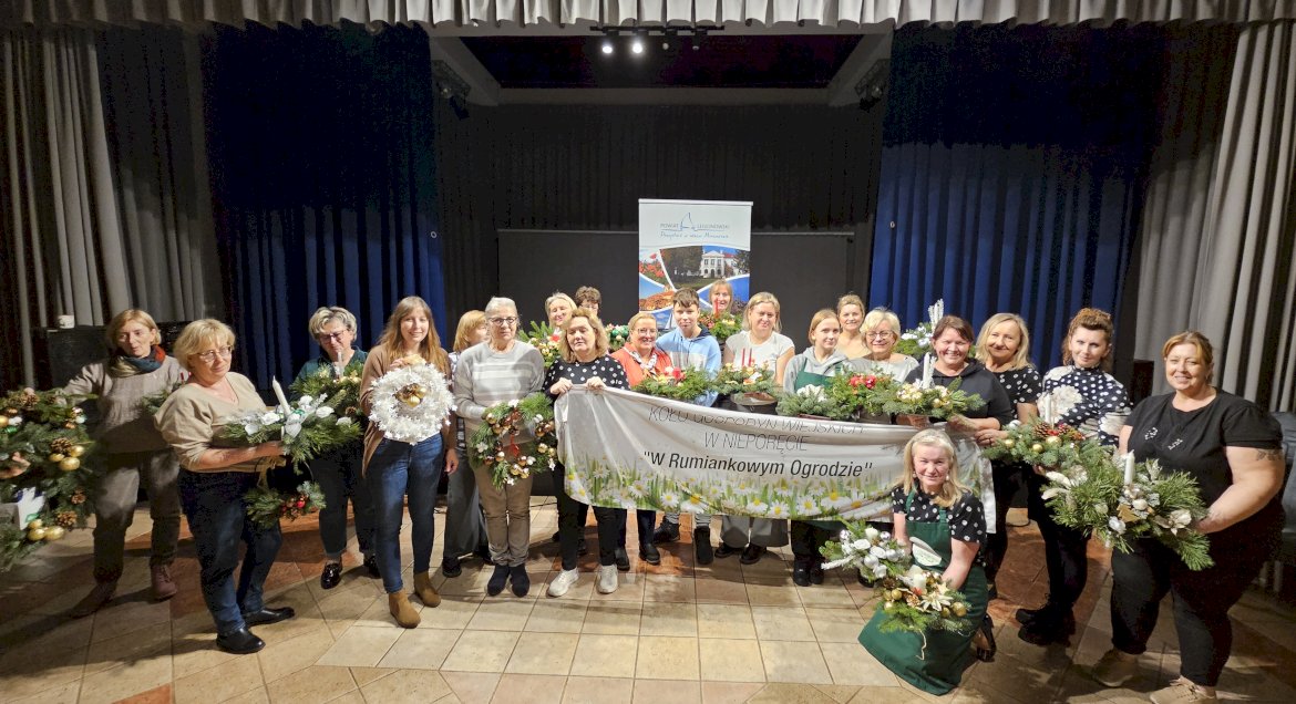 Zdjęcie przedstawia grupę osób ze stroikami w dłoniach, uczestników warsztatów stroikowych w tle rollup Powiatu Legionowskiego