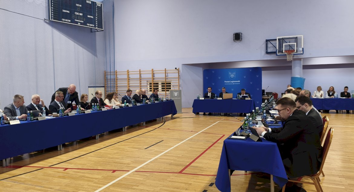 Widok sali, w której centralnie ustawiono stół prezydialny oraz dwa długie stoły ustawione prostopadle do niego. Przy stołach siedzą radni Rady Powiatu w Legionowie - podczas obrad sesji Rady Powiatu.