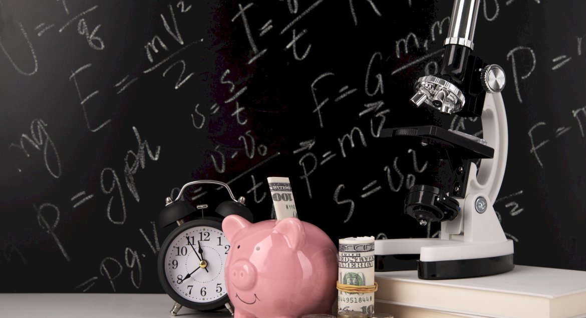 Na zdjęciu czarna tablica kredowa, a przed nią skarbonka świnka, zwinięty rulon pieniędzy, zegarek i mokroskop