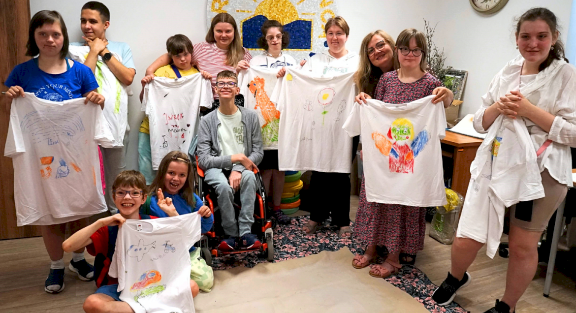 Na zdjęciu grupowym znajdują się podopieczni Fundacji Promień Słońca, wszyscy trzymają w rękach białe koszulki z kolorowymi rysunkami wykonanymi własnoręcznie