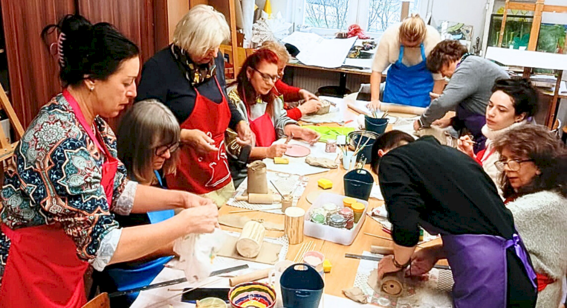 Na zdjęciu widać jak uczestniczki warsztatów ubrane w fartuchy siedzą a część stoi przy stole, na którym znajduje się glina, farby i wiele przyrządów do wykonywania talerzy ceramicznych