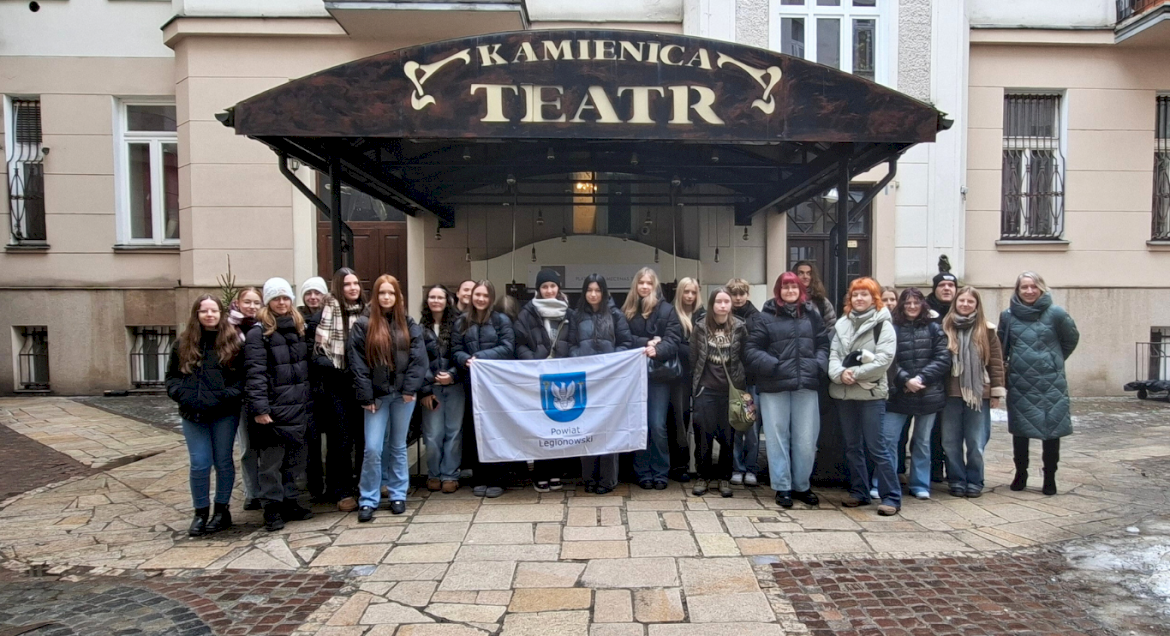 Na zdjęciu widać grupę młodzieży z opiekunami stojący przed Teatrem Kamienica z flagą Powiatu Legionowskiego